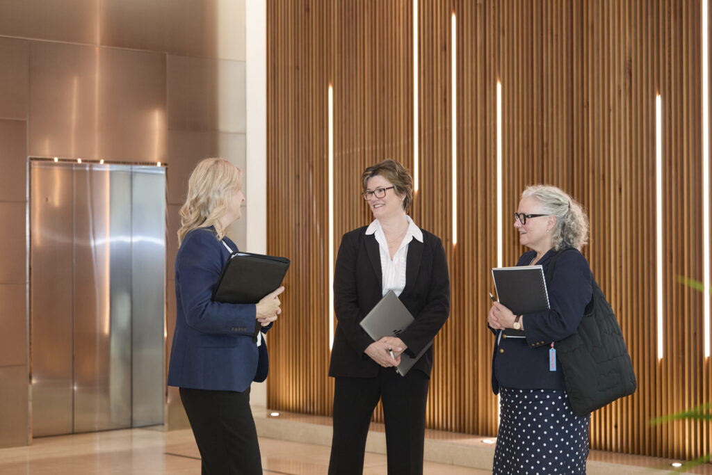 Three women in business attire talk by a wood-paneled wall in a modern lobby, possibly discussing a lawyer's case.