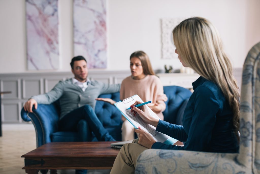 A couple discusses Family Dispute Resolution with a therapist taking notes on a clipboard in a bright, comfortable room.