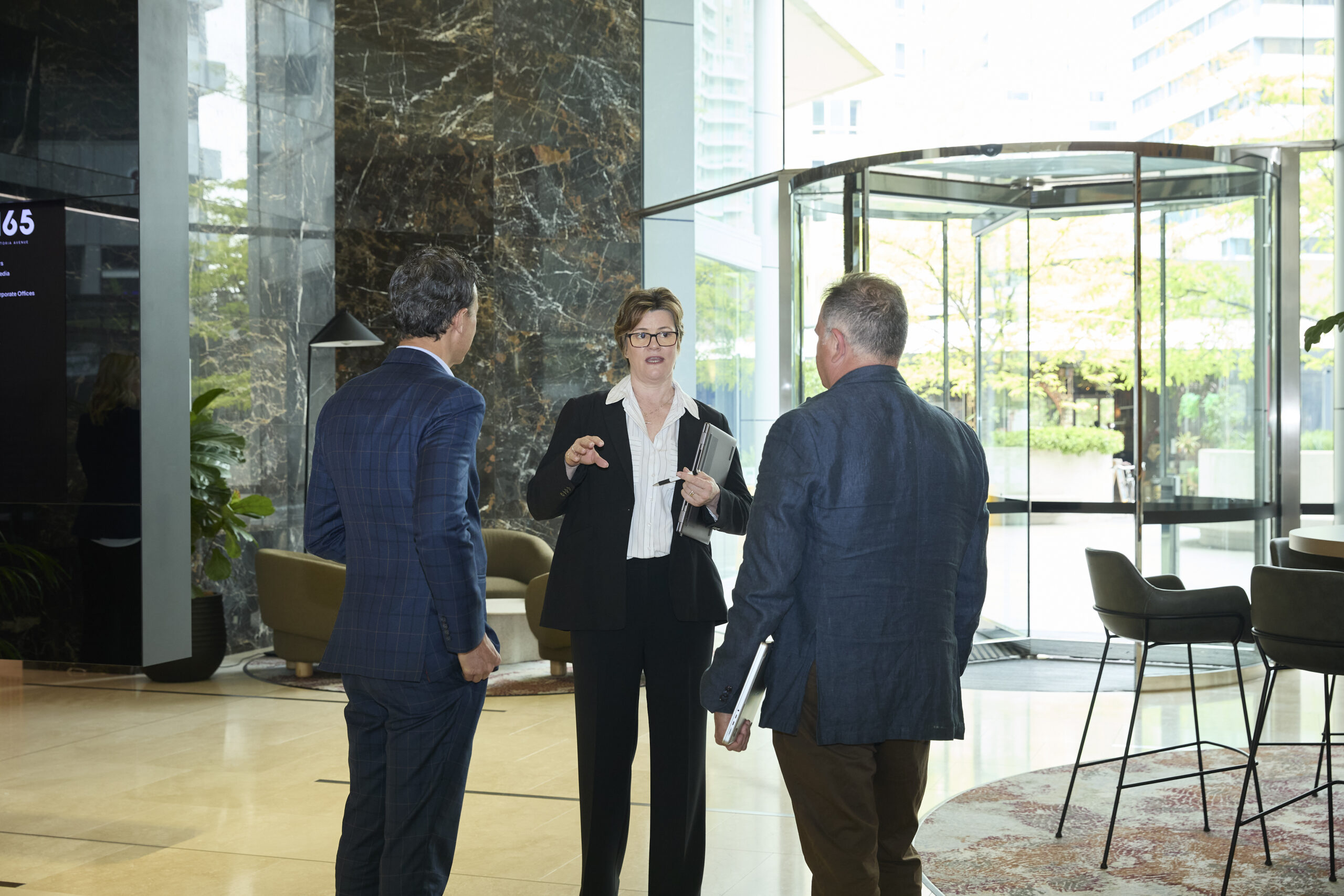 Three professionals discuss Family Dispute Resolution in a modern lobby near a glass revolving door, business attire.