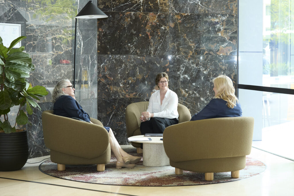 Three women sit in armchairs around a table, ready for mediation in a modern lobby with marble walls and large window.