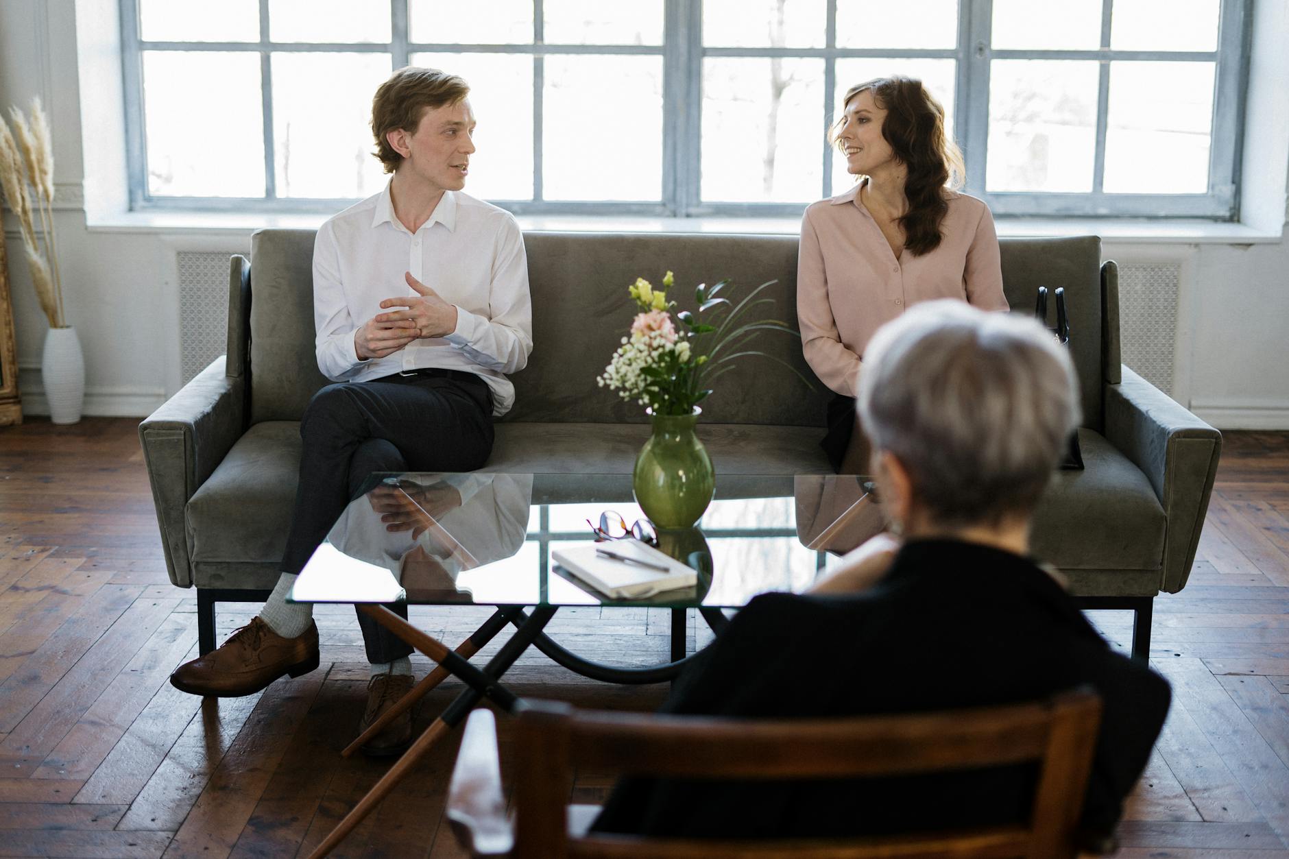 A man and woman sit on a sofa in a therapy or counseling session, facing a gray-haired therapist with certificates and notebook.