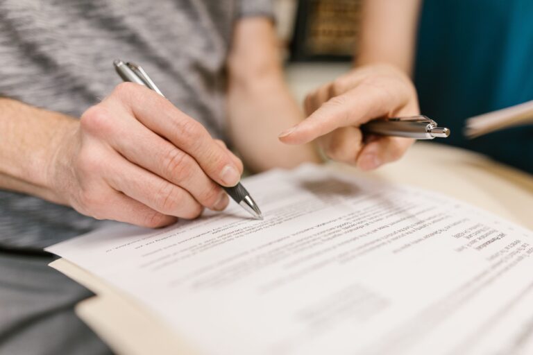 Two hands sign a document with a black pen while a second hand holds a silver pen nearby, on a desk surface.